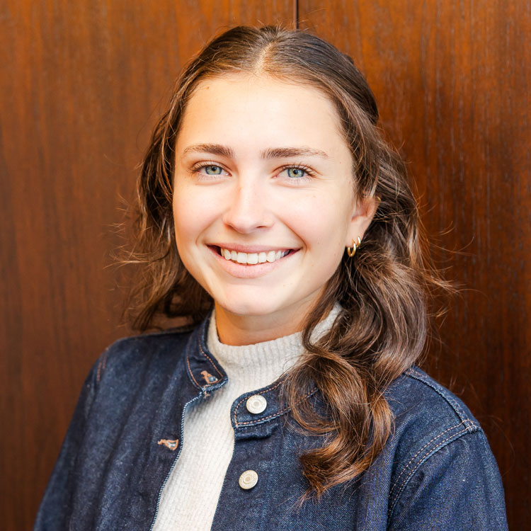 Reagan M. Petrow Reagan M. Petrow: Smiling person with shoulder-length brown hair, wearing a denim jacket over a light sweater. Background is a wooden panel, conveying a warm tone.