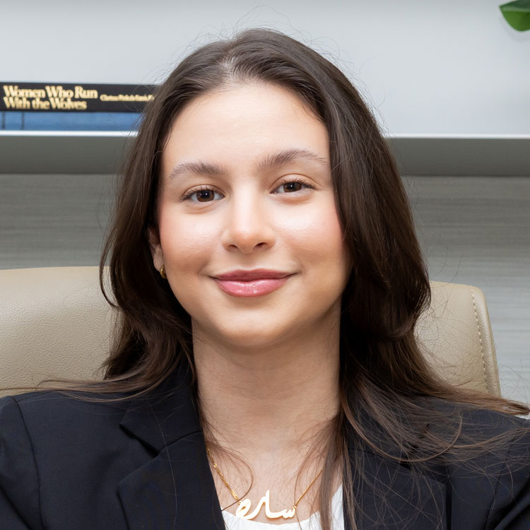 Sara Nasif | Professional portrait of a young woman in a black blazer, seated in an office chair with a neutral smile and a gold necklace.