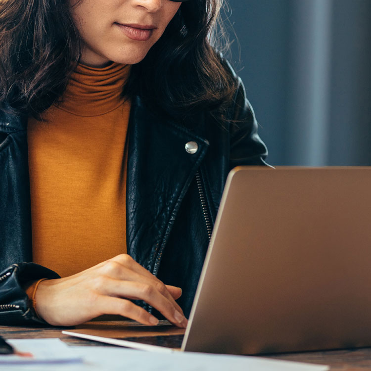 Resources | Important Links: A woman in a leather jacket and mustard sweater focuses on her laptop, working diligently. Soft lighting and a blurred background create a calm atmosphere.