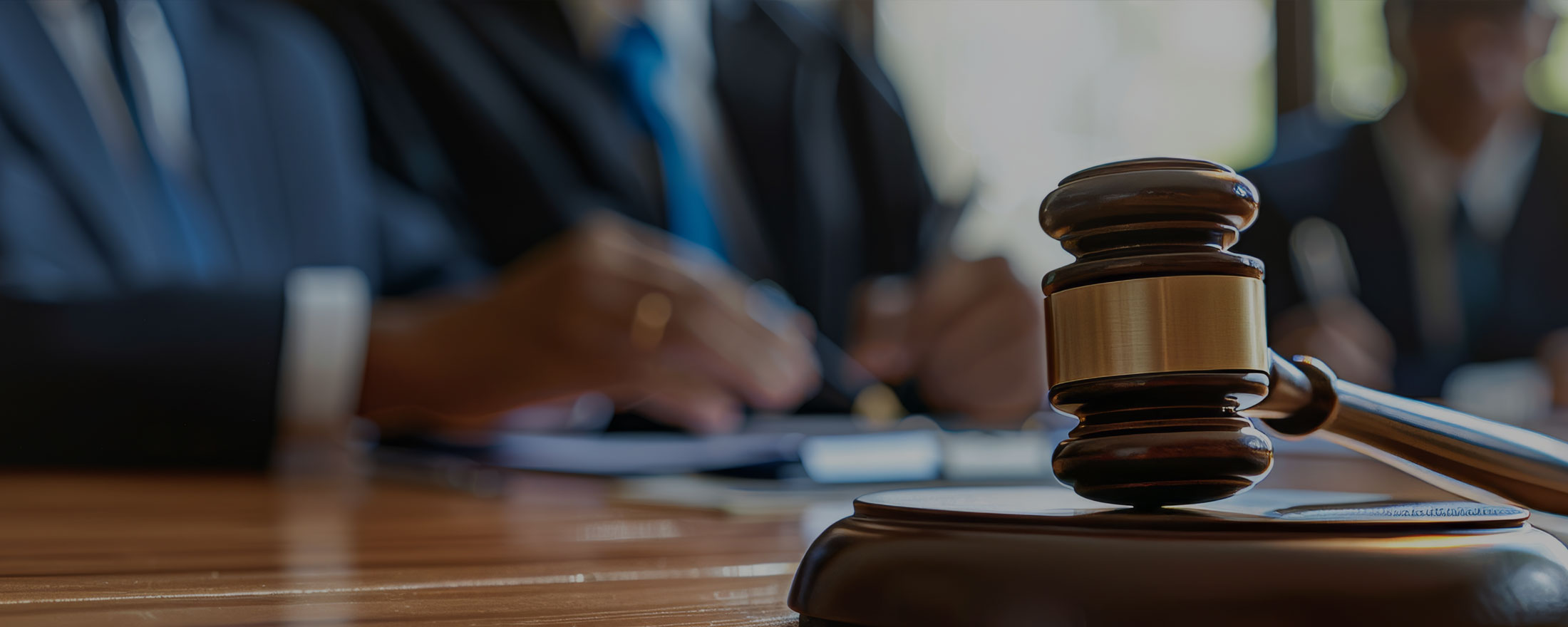 Law Better: Our Team | Gavel on a sound block on a courtroom desk with blurred lawyers in suits in the background of the scene.